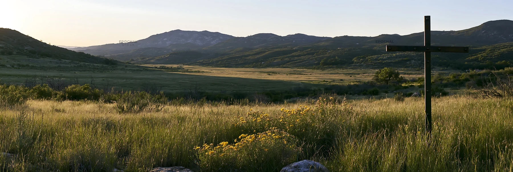 Iron Christian cross overlooking valley meadow at sunset - AI-generated stock image