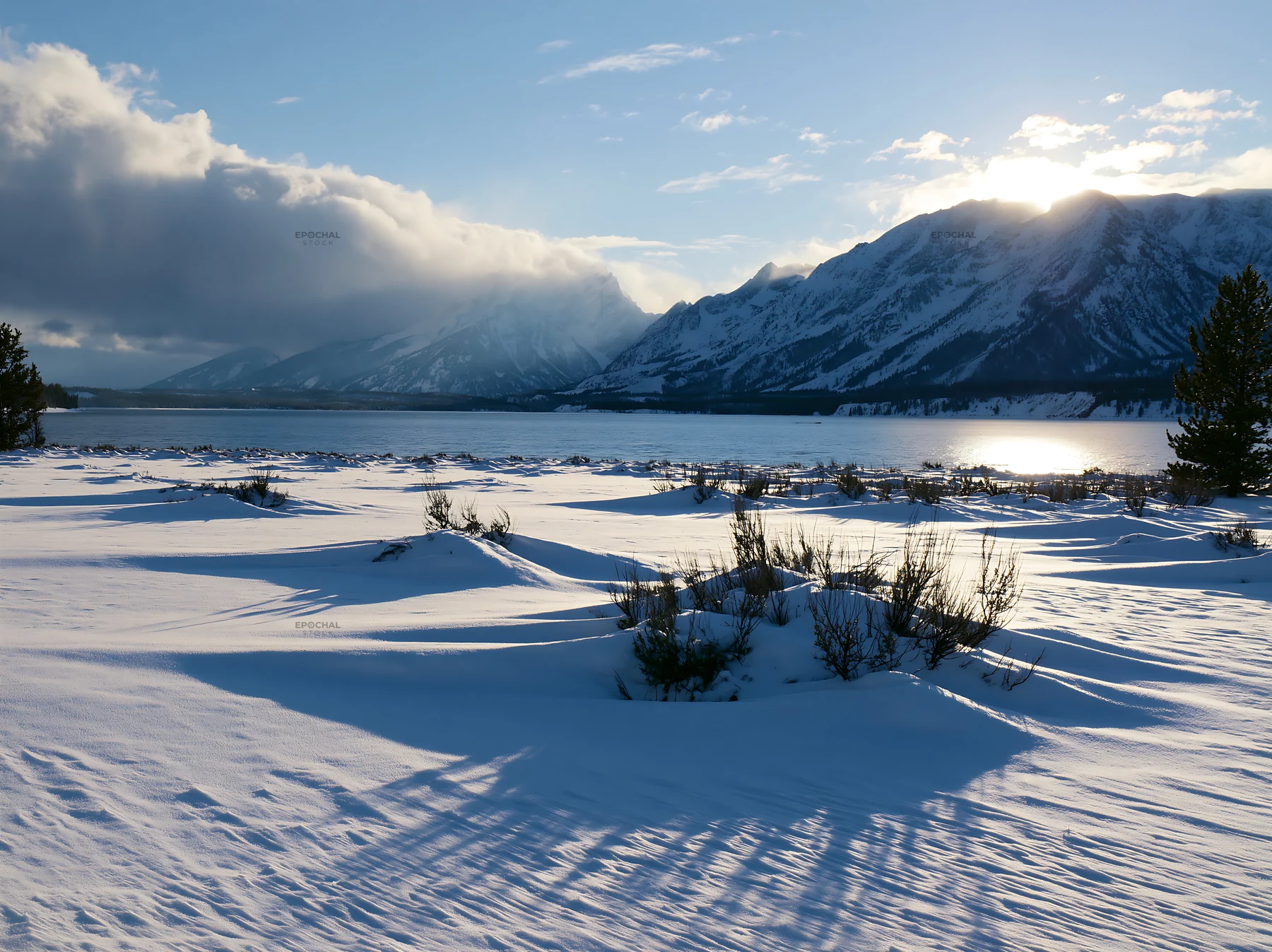 Snowy lakeside meadow with mountains and sun in the background - AI-generated stock image