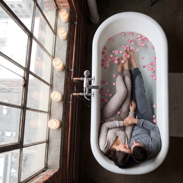 Couple relaxes in bathtub with rose petals near large window - stock photo