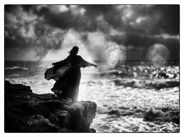 Woman stands on cliff by ocean during cloudy weather at sunset - stock photo