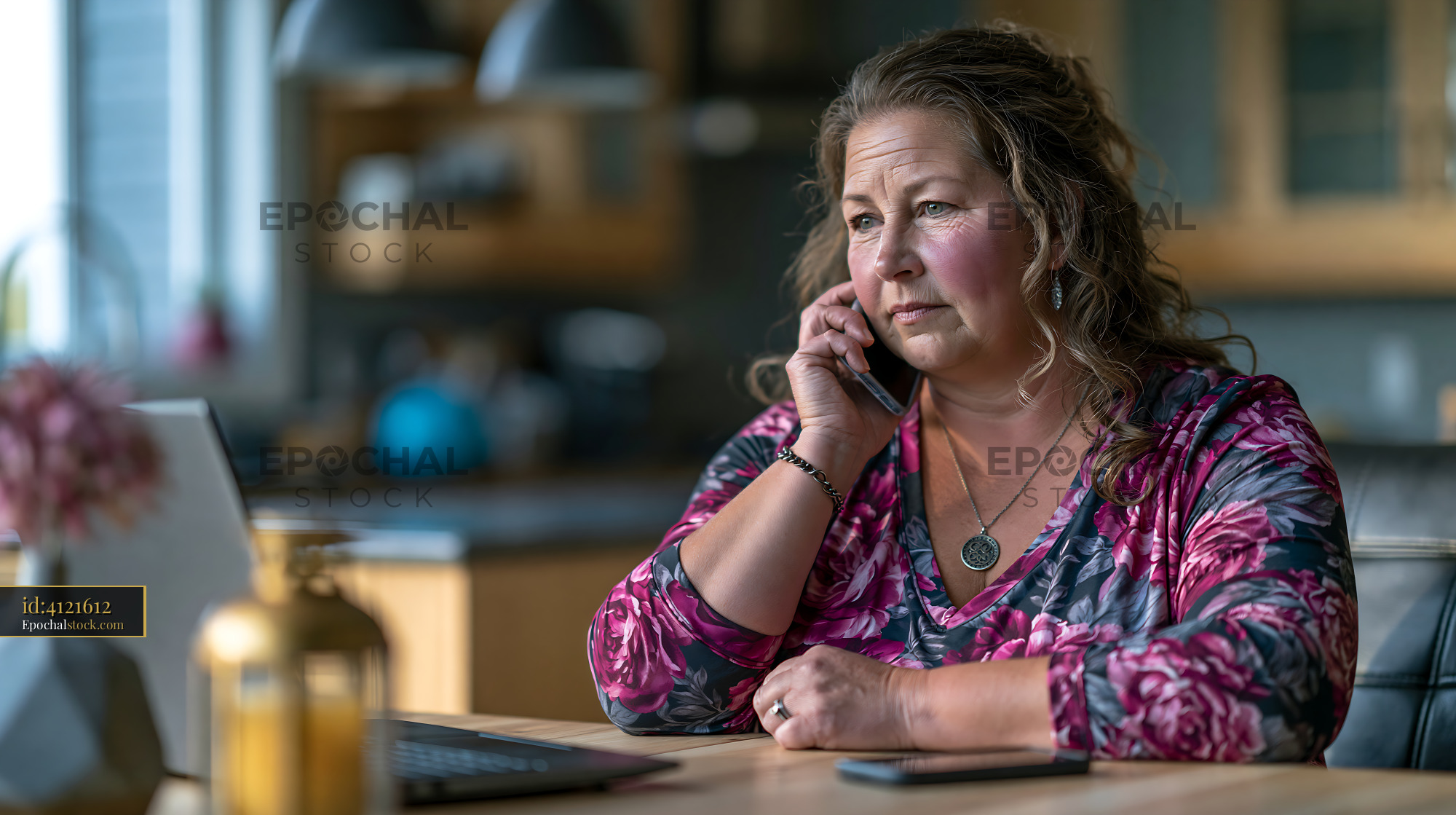 Watermarked full-resolution preview of Woman talking on the phone while sitting at a table with a laptop