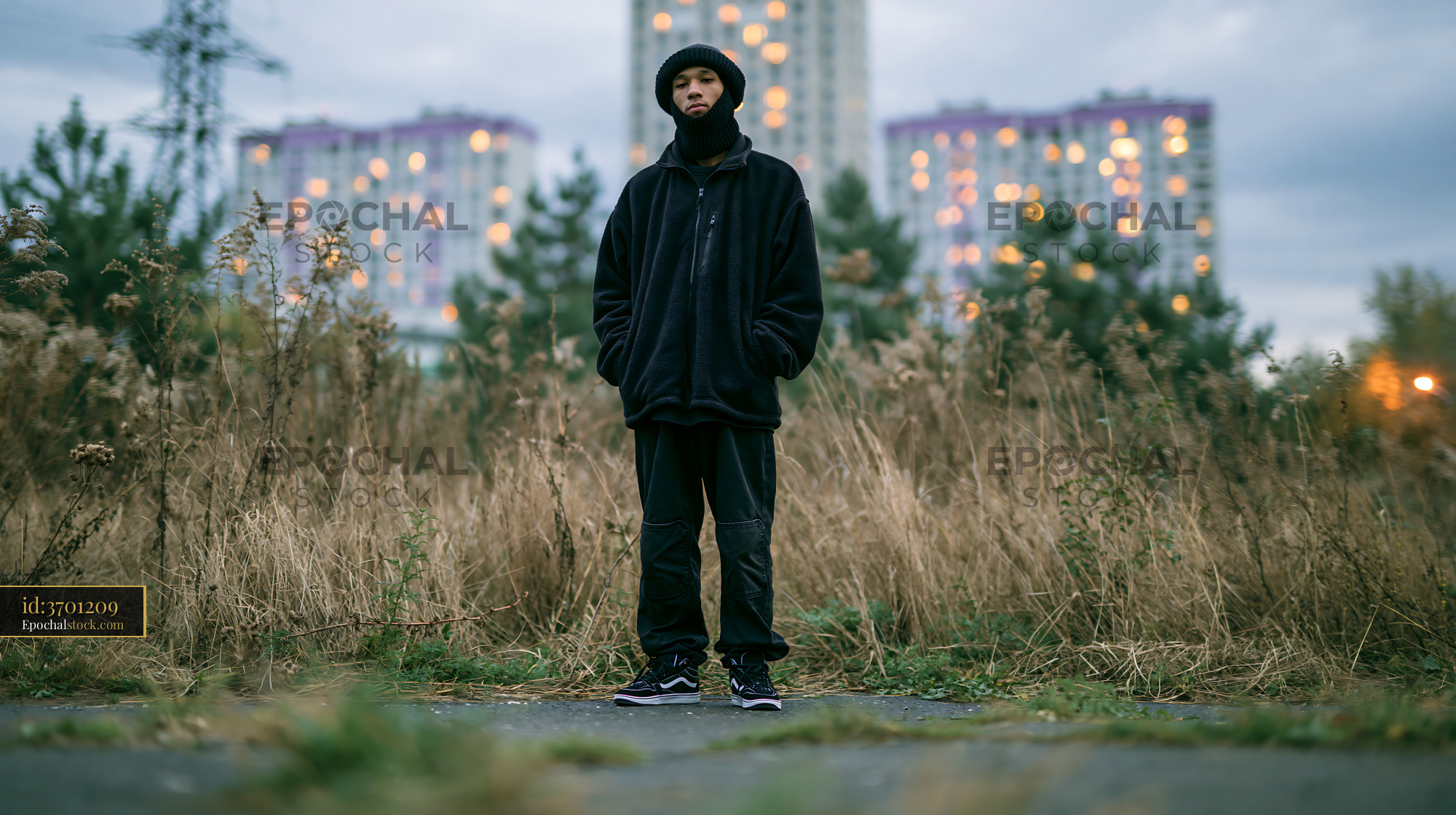 Watermarked full-resolution preview of Person standing in tall grass near buildings at dusk