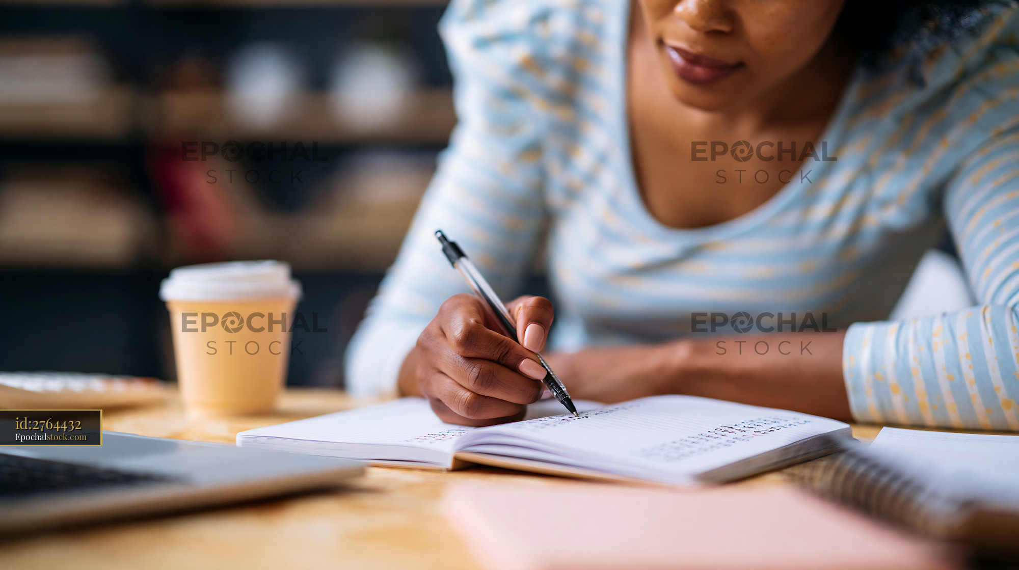 Watermarked full-resolution preview of Woman writing notes in a notebook at a wooden table with a laptop