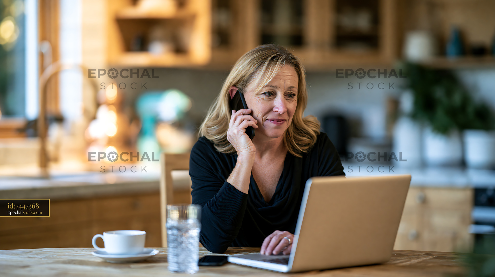 Watermarked full-resolution preview of Woman using laptop while talking on phone in kitchen