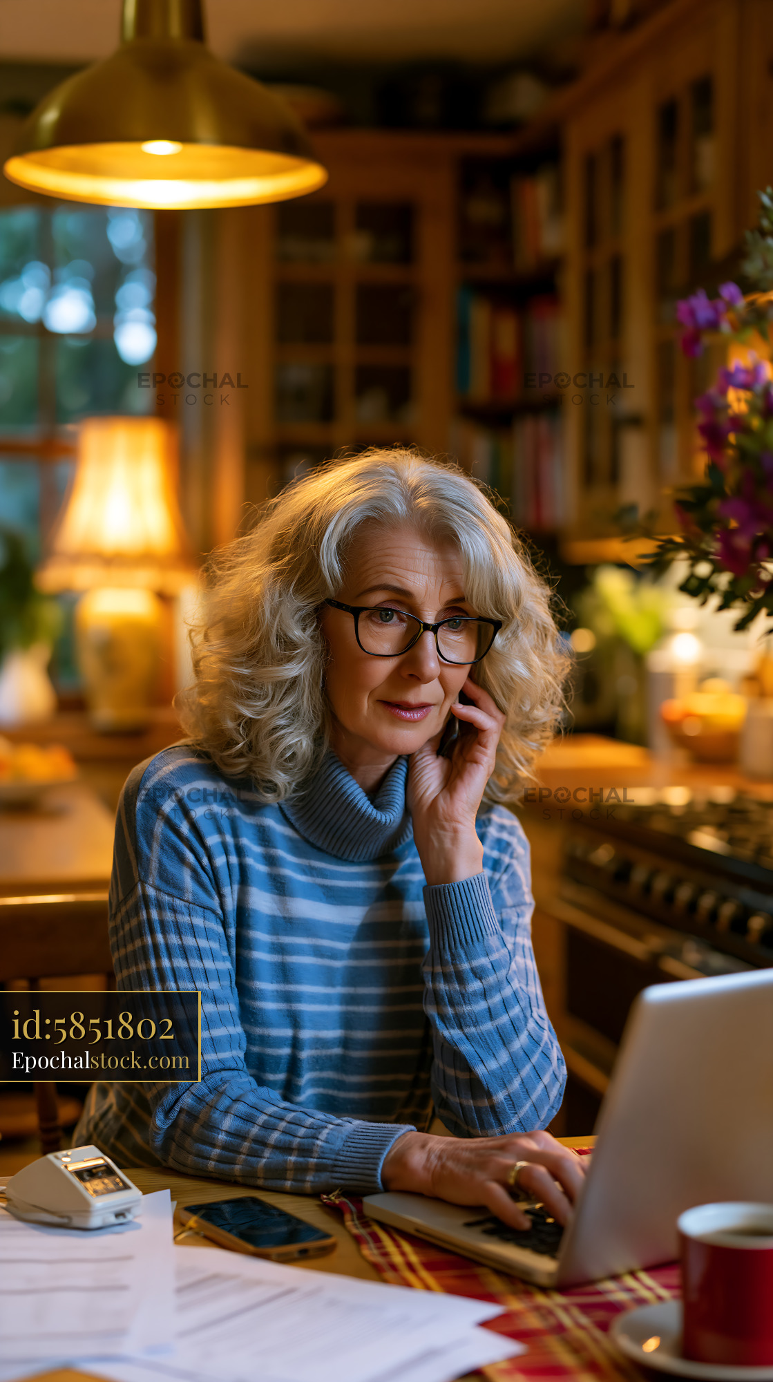 Watermarked full-resolution preview of Older woman on phone while using laptop in cozy kitchen