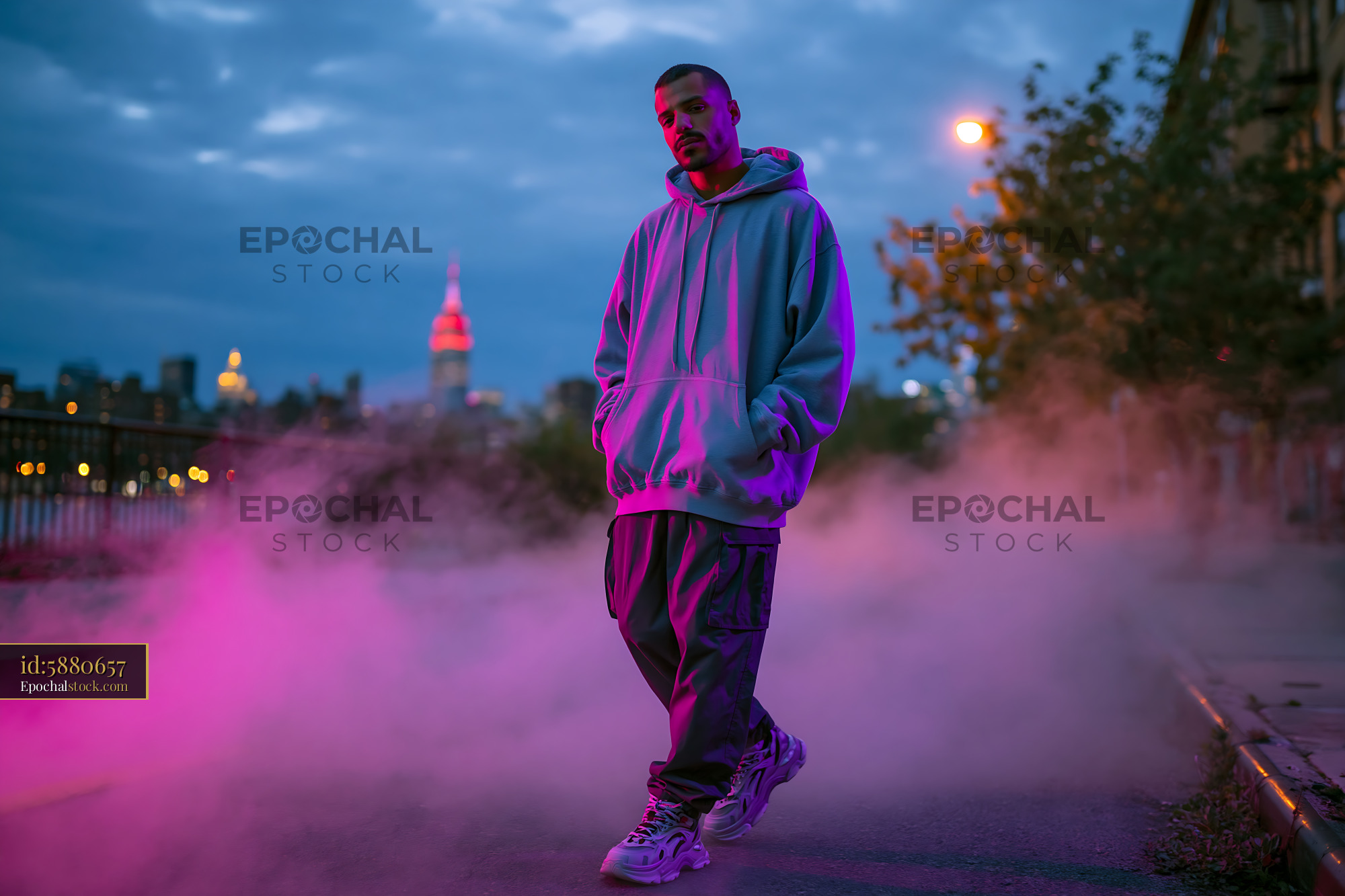 Watermarked full-resolution preview of Man in gray hoodie stands on street with city skyline at dusk