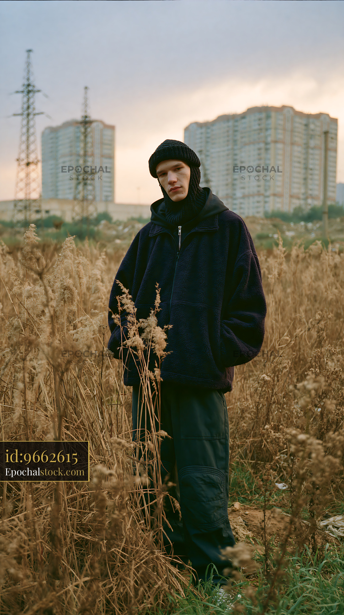 Watermarked full-resolution preview of Young person standing in tall grass near buildings during sunset