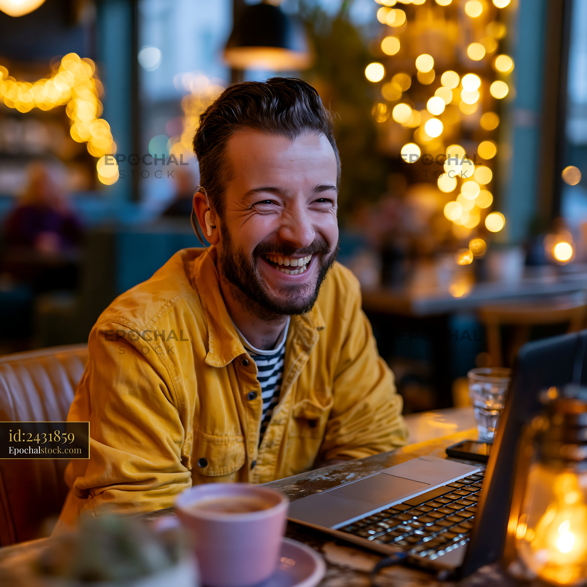Watermarked full-resolution preview of Man smiling while working on a laptop in a cozy cafe with lights