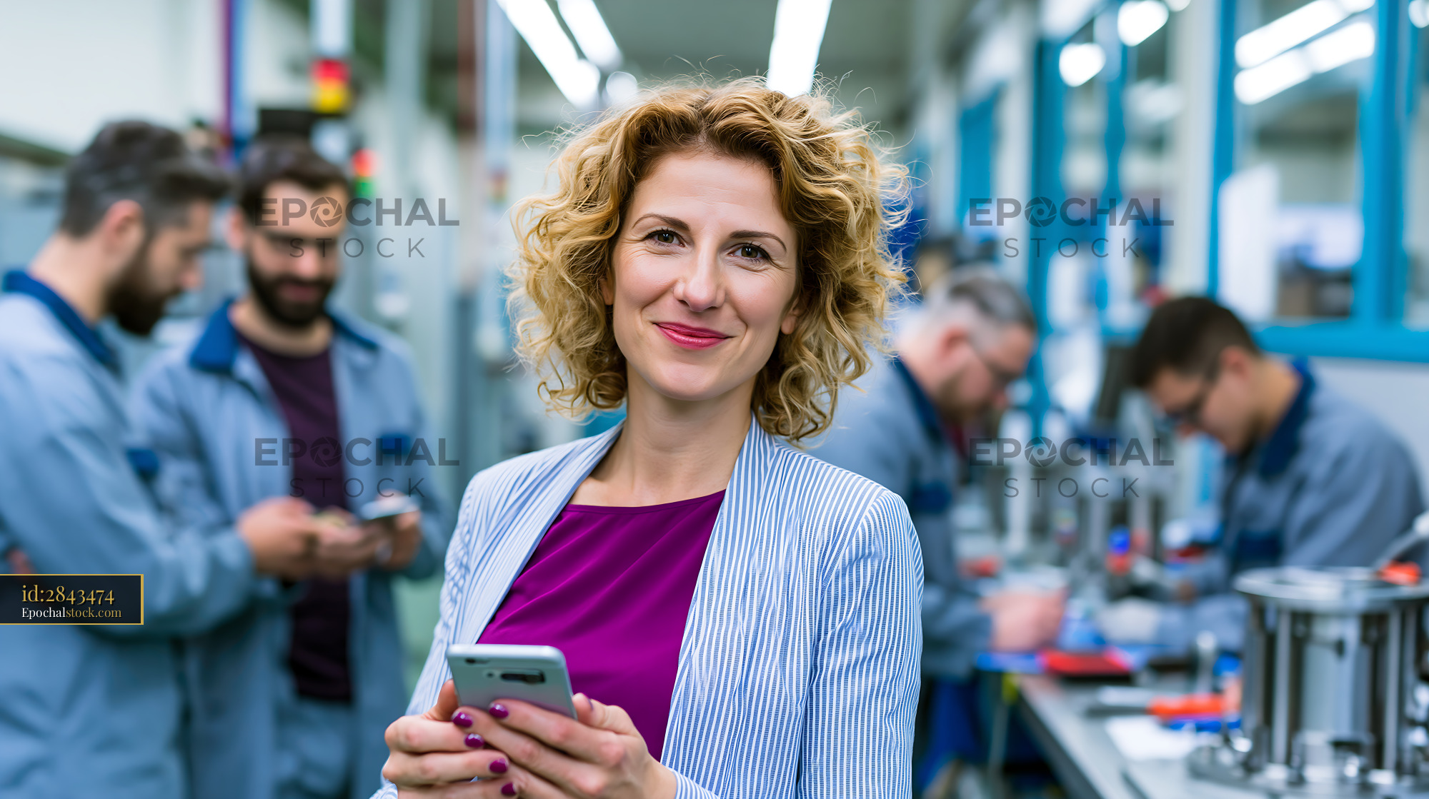 Watermarked full-resolution preview of Woman holding smartphone in a factory with workers in the background