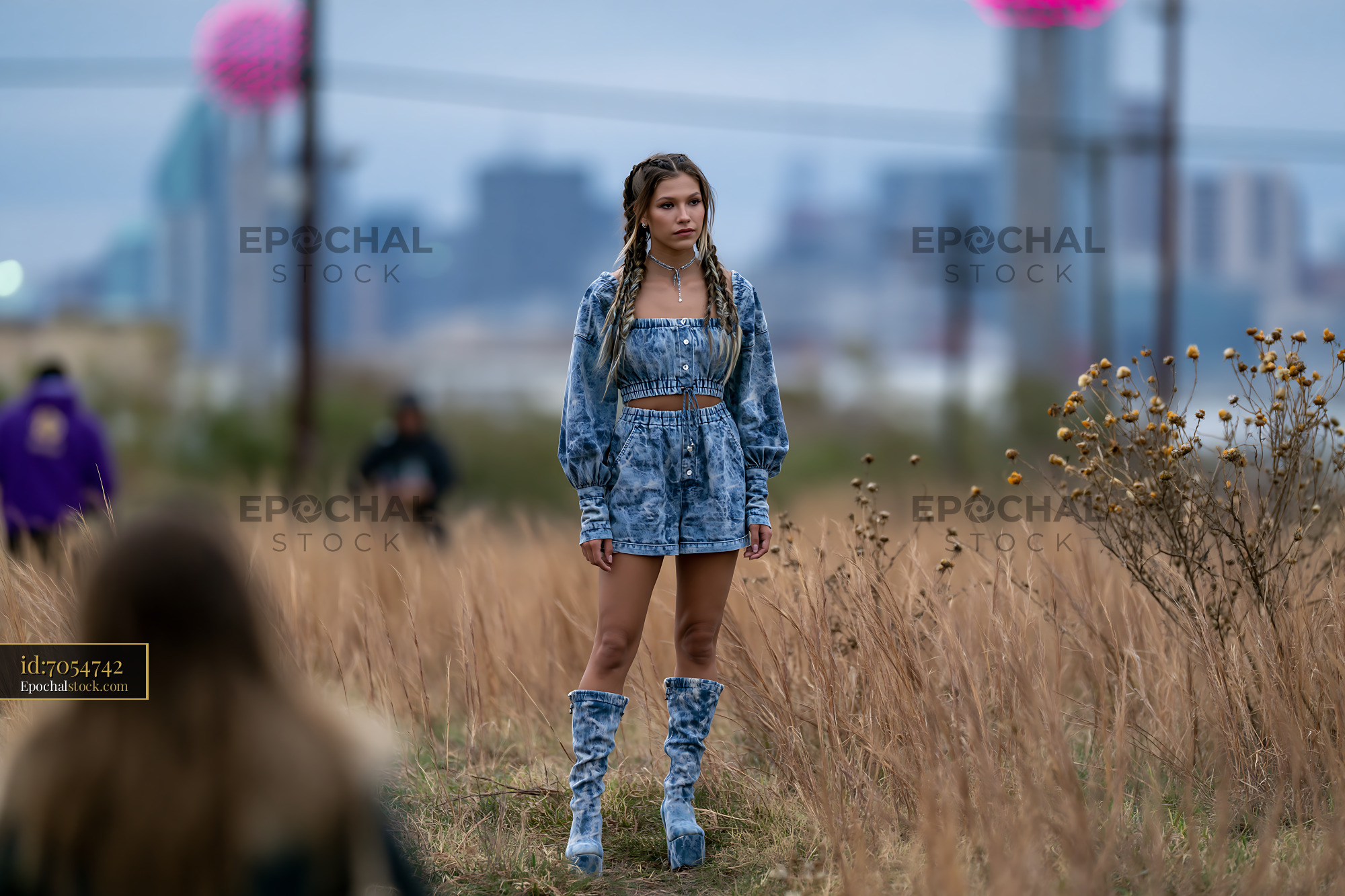 Watermarked full-resolution preview of Young woman in denim outfit standing in a field with city skyline