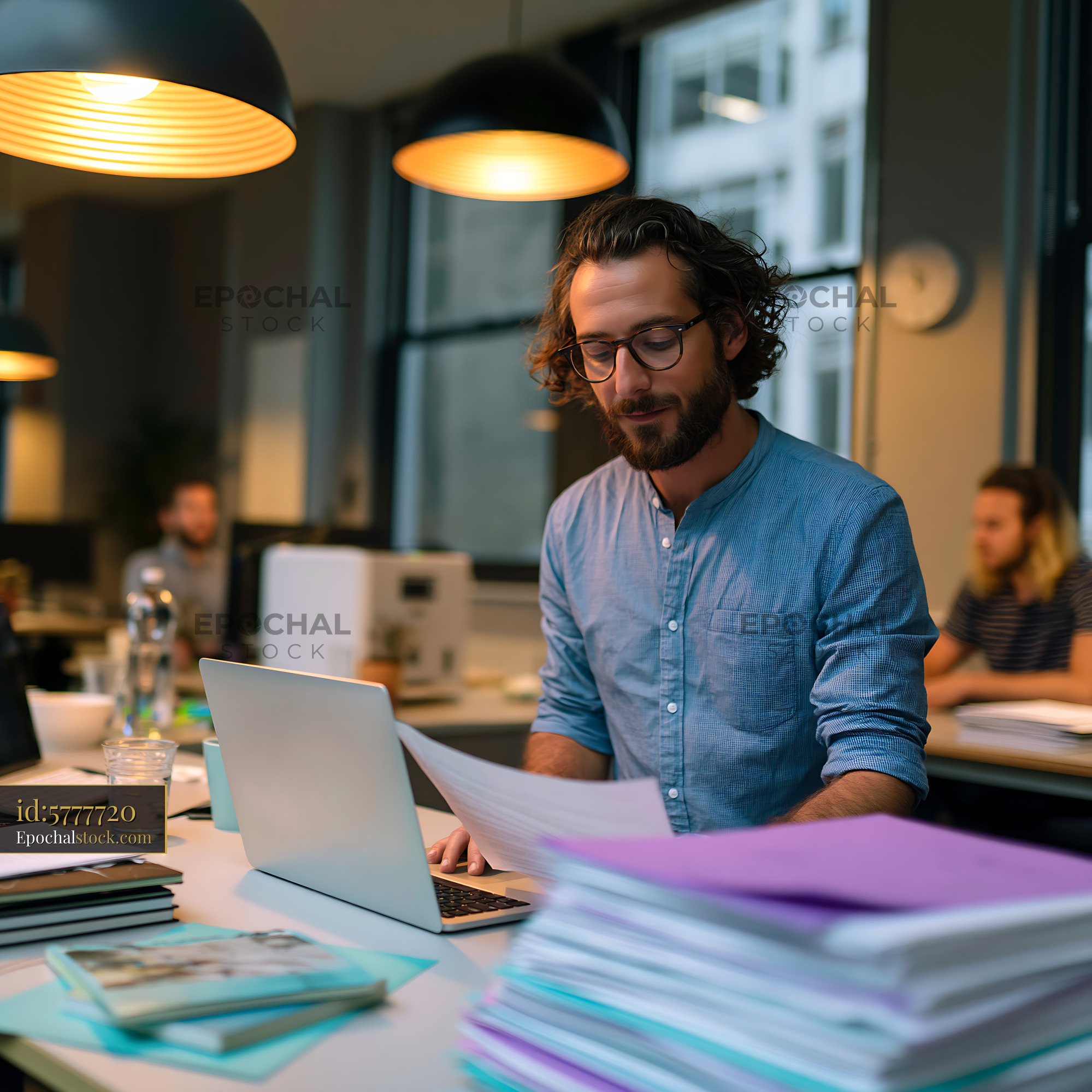 Watermarked full-resolution preview of Man working in a modern office with paperwork and a laptop