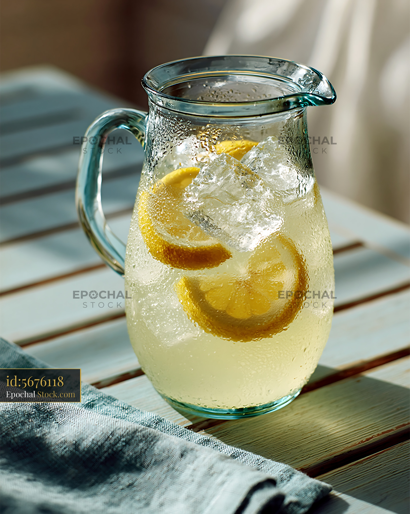 Glass lemonade pitcher with ice cubes and lemon slices on sunny table - stock photo