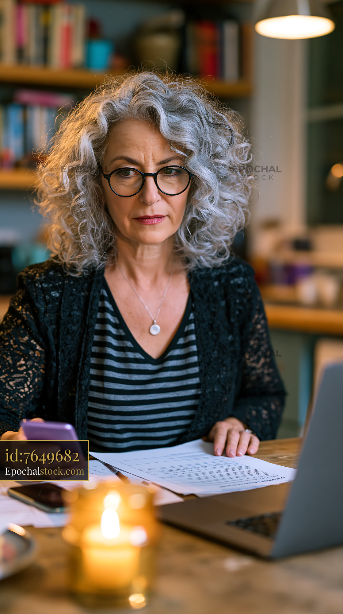 Watermarked full-resolution preview of Woman working on documents at a table with a laptop and phone