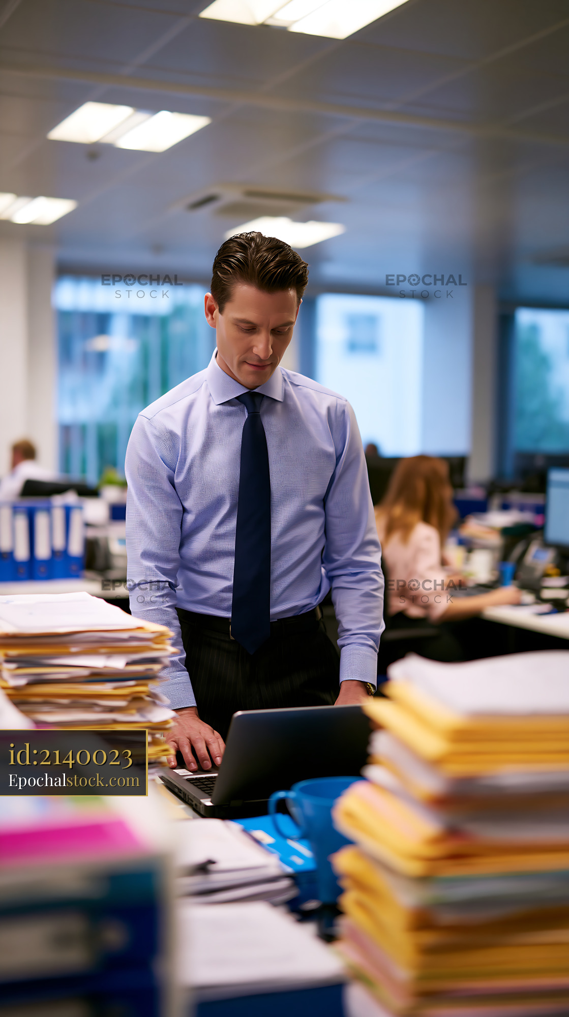 Watermarked full-resolution preview of Office worker standing at desk with paperwork in busy environment
