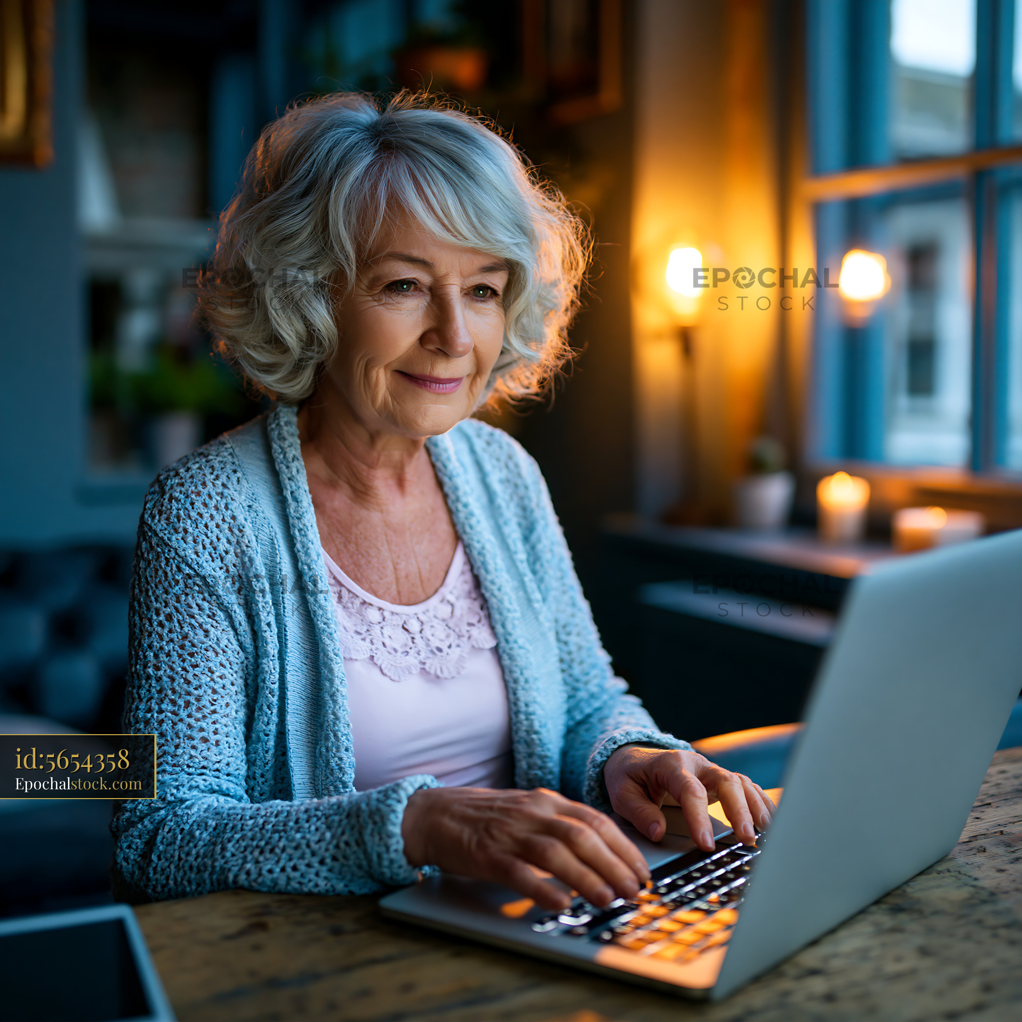 Watermarked full-resolution preview of Senior woman working on laptop at home in cozy evening light