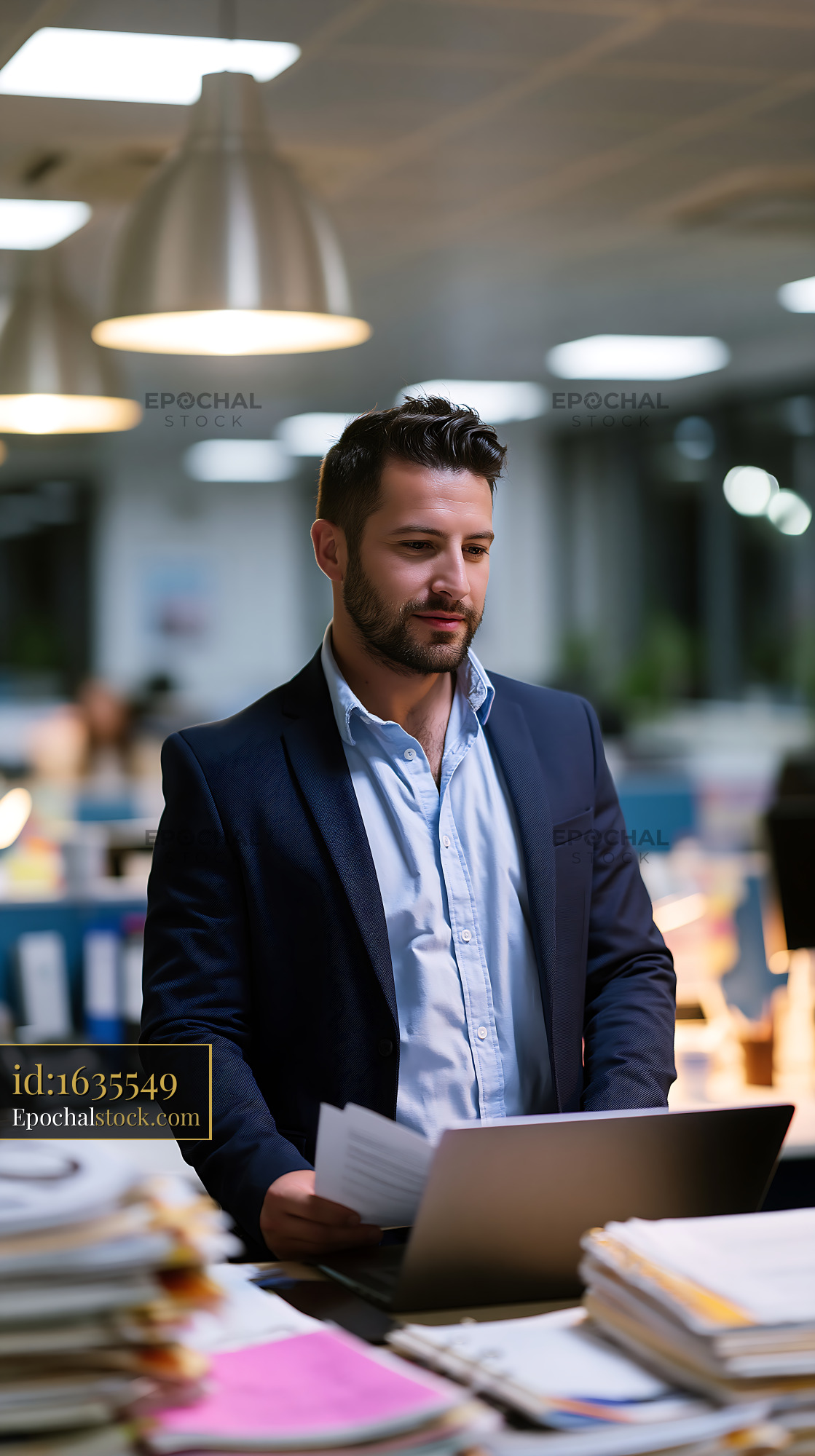 Watermarked full-resolution preview of Man stands at desk with laptop in busy office setting