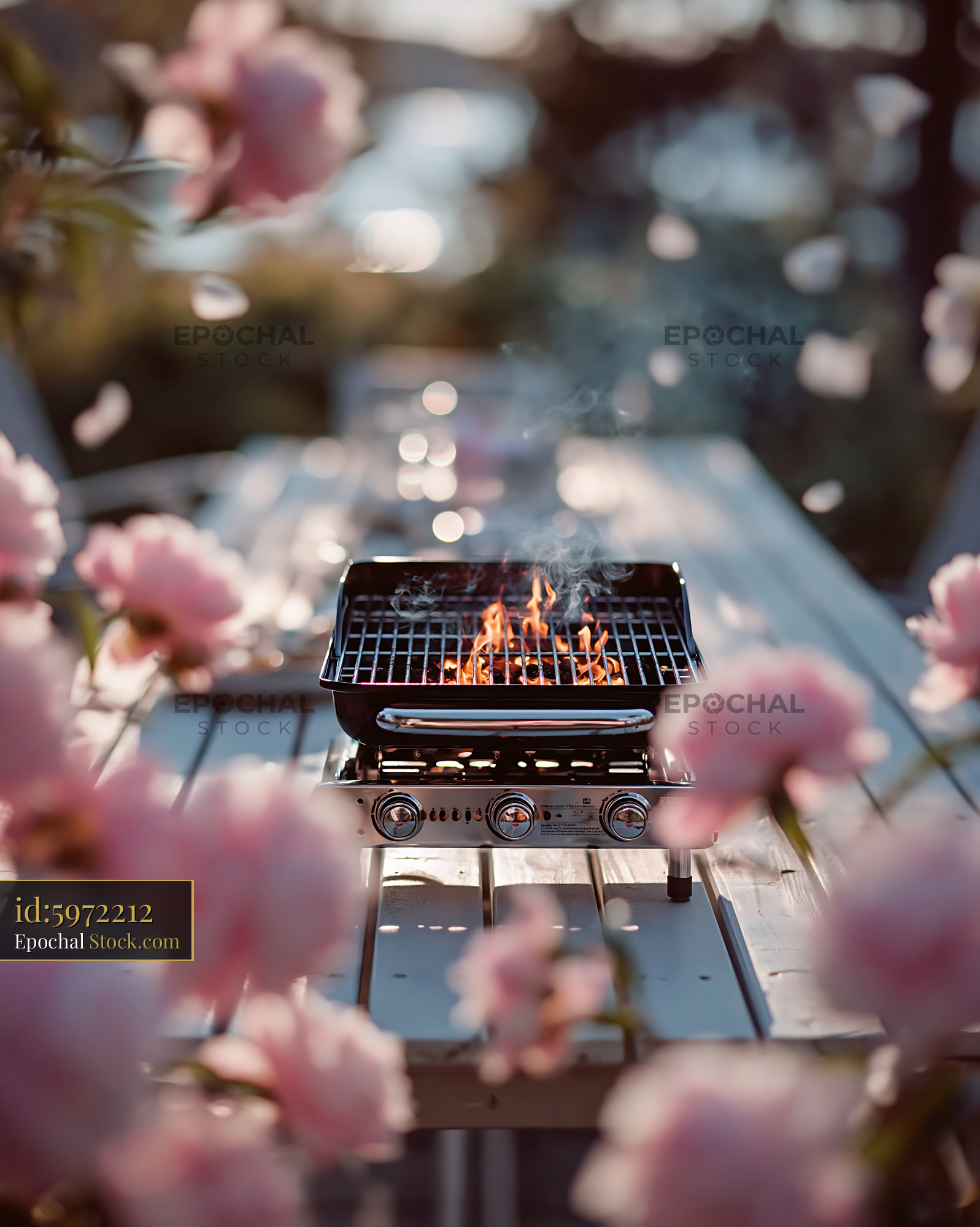 Portable gas grill with flames on a garden table among pink flowers - stock photo